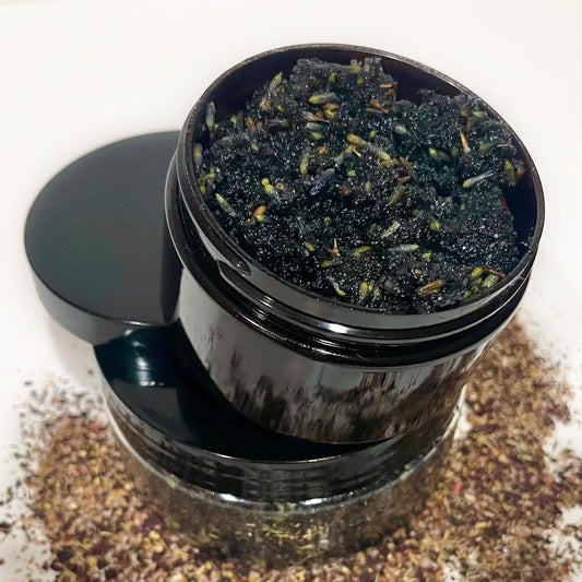 Two containers of the product, Black Velvet Scub are stacked on top of each other with lavender petals scattered around the products.